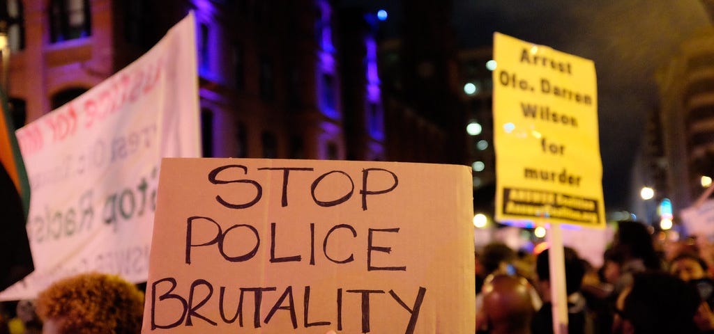 Image of a nighttime protest with a cardboard protest sign in the center. The sign says “Stop Police Brutality” in all caps.