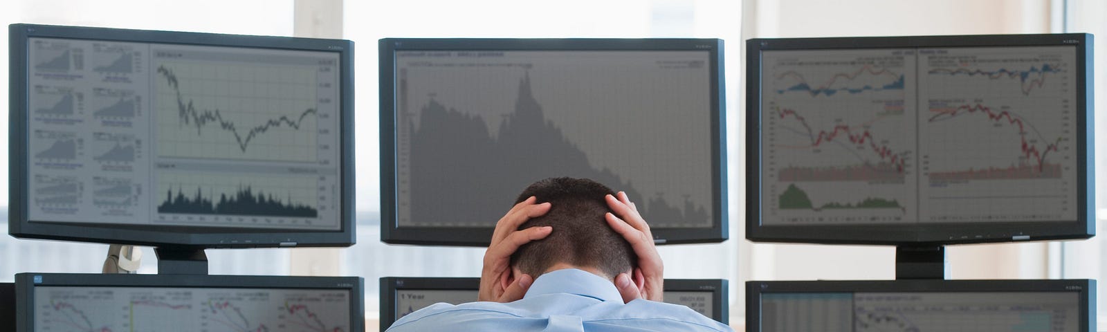 A frustrated worker with their hands on their head sits down at their desk, which has six computer monitors.