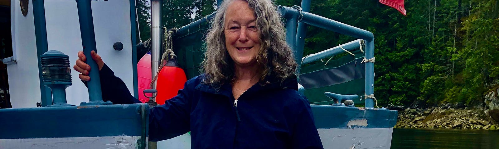 A woman stands on the stern of a boat. She is holding a rope and holding on to the boat with the other. She has long grey hair.