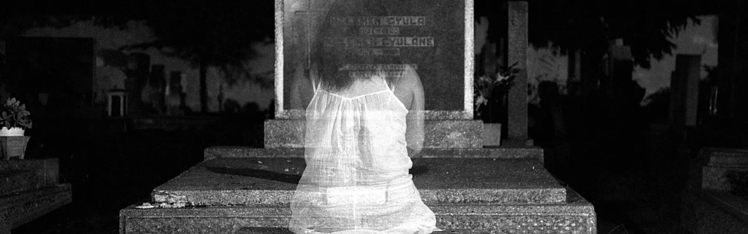 A young woman, wearing a white nightie, is a shadow of herself as she sits in shock, reading her name engraved on her own tombstone. She is in fact a ghost.