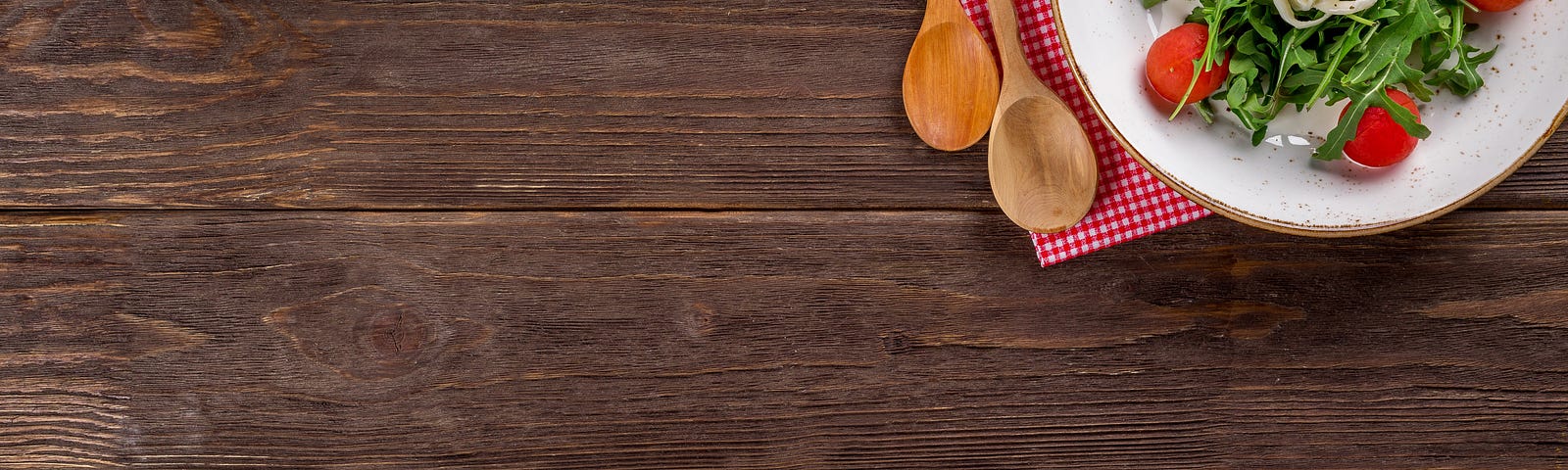 A wooden table top with two wooden spoons, a red checked napkin, and a bowl of salad