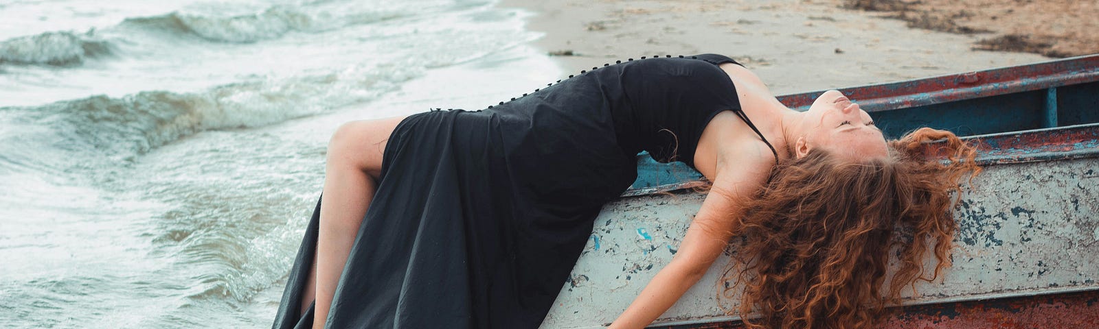 Young woman with long red hair, wearing a long black dress, draped over rowboat as the ocean washes over her feet.