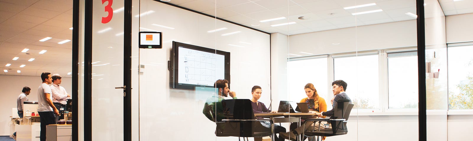 Four people in discussion while sitting at a meeting table in a glass-walled meeting room.