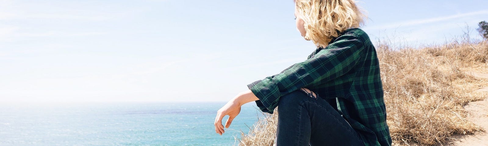 blond woman sitting on a cliff overlooking the ocean imagining her dreams coming true