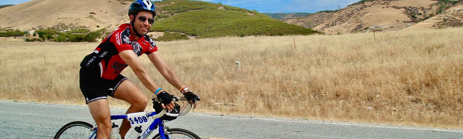 A young man, the author, sits atop a roadbike and wears a helmet as he rides along a paved road with a dry-grass backdrop.