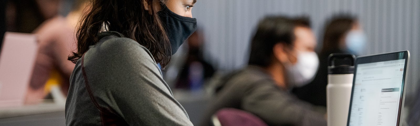 A woman wears a face mask while typing on her laptop. She is surrounded by other masked students in a lecture hall.