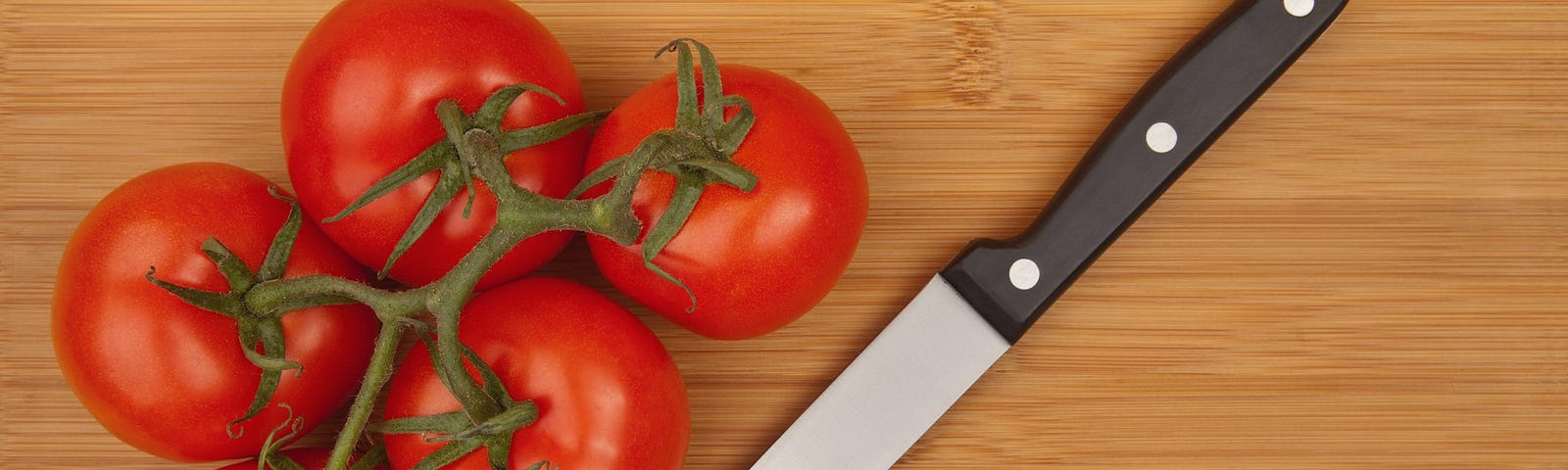 Five ripe tomatoes still connected on the vine sitting on a wooden cutting board next to a kitchen knife with a black handle