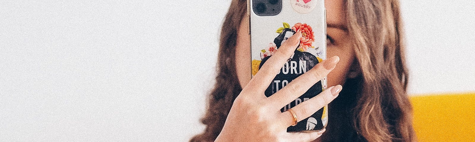 White woman with long brown hair, wearing a white shirt and jeans, takes a selfie in the mirror with the camera covering her face.