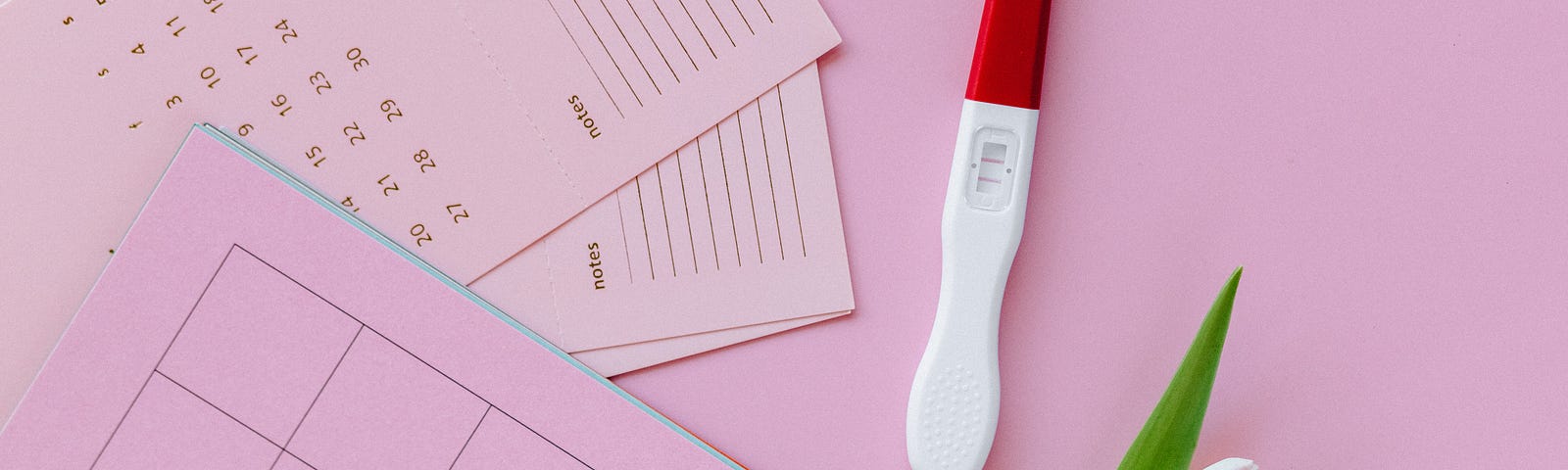 Red and white pregnancy test lying on pink stationary next to a pink tulip