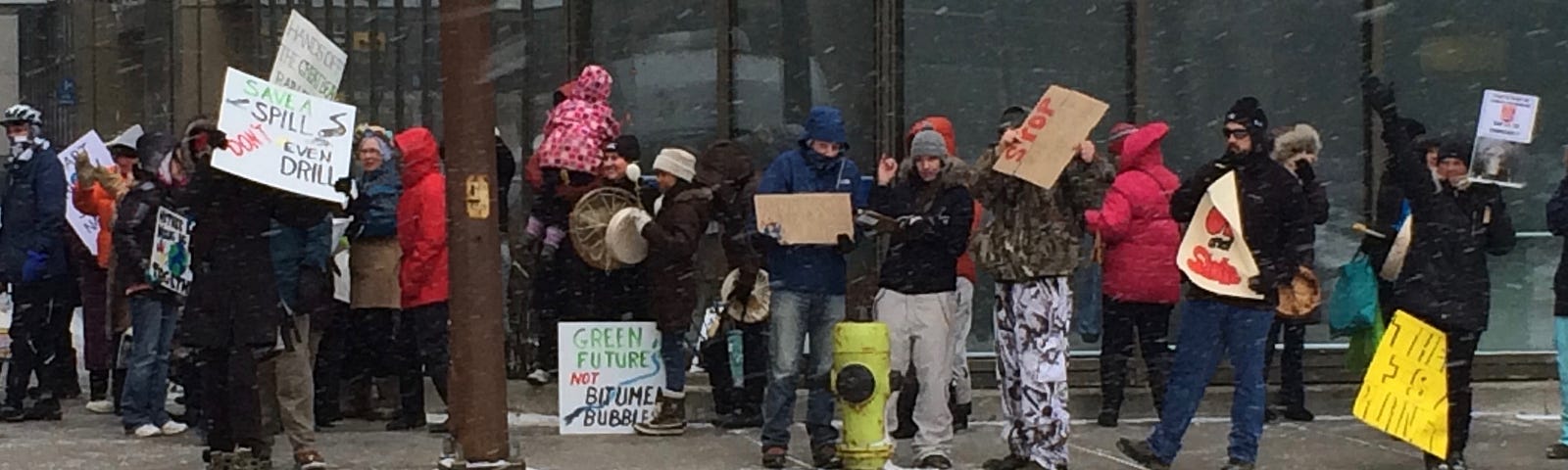Protestors along a sidewalk, mostly dressed in winter garb, with placard signs and drums, while snow is falling.
