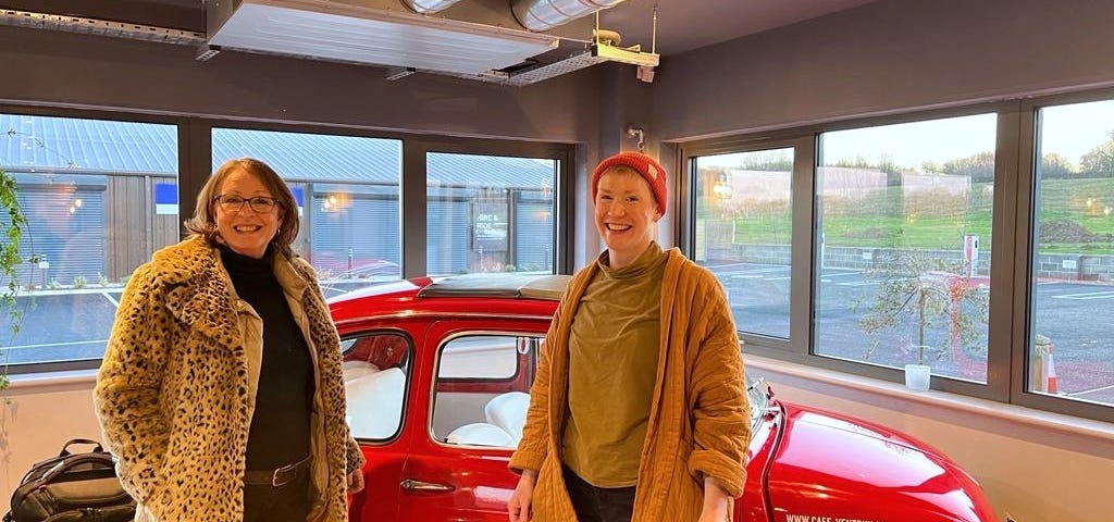 Two White women smiling and standing in front of a red vintage car in a cafe