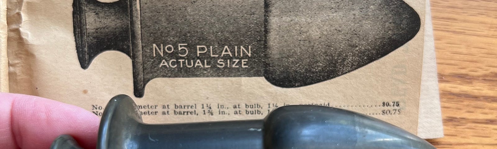 In the foreground, a left hand holding a vintage, plastic “rectal dilator.” In the background, an etching of the same dilator, labeled “№ 5 PLAIN ACTUAL SIZE.”