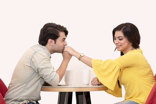 Man and woman at a table, cups of coffee, he kisses her hand, she smiles but looks away