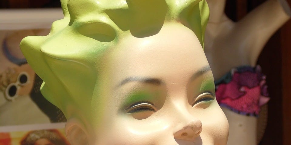 Head of mannequin with green hair and eye shadow, with red lips in a wide- open smile.