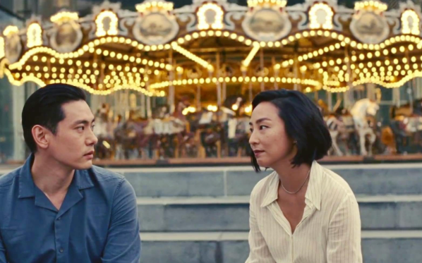 A Korean man and a Korean woman sit in front of a carousel. They gaze longingly into each other’s eyes. Film review Past Lives, critic, Oscars Best Picture, Academy awards, writing, script, drama, comedy, award winning films, film review critic