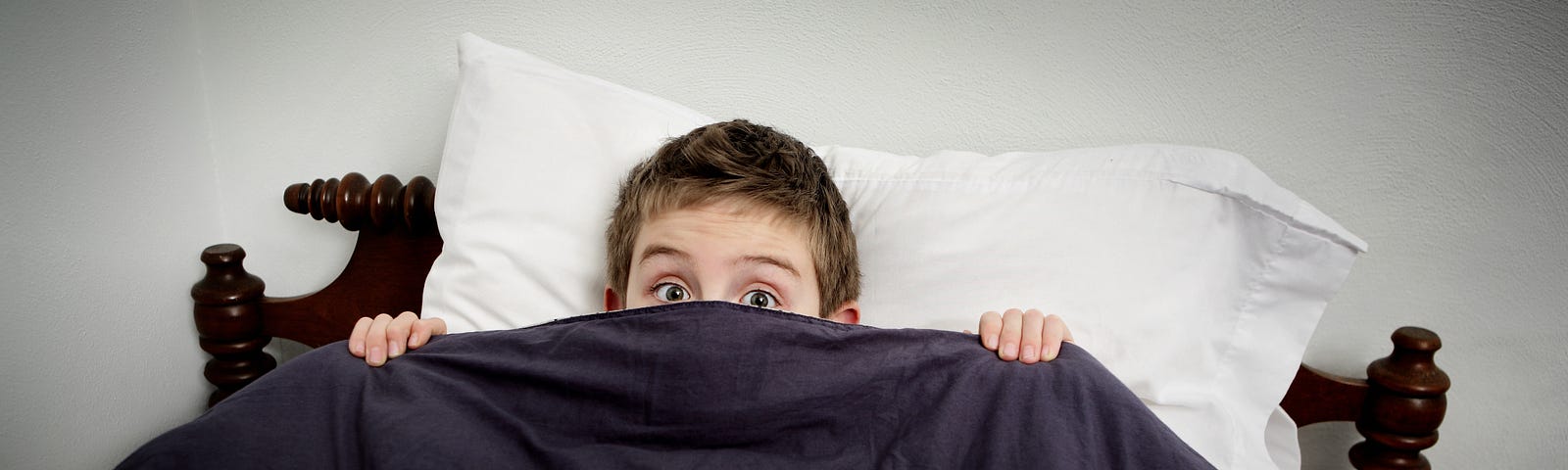 Young boy in bed, peeking over his blanket in fear