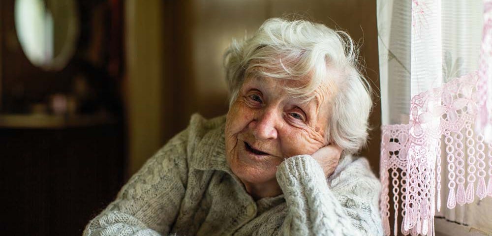 What’s too old? Elder woman smiling at the camera while sitting next to a window.