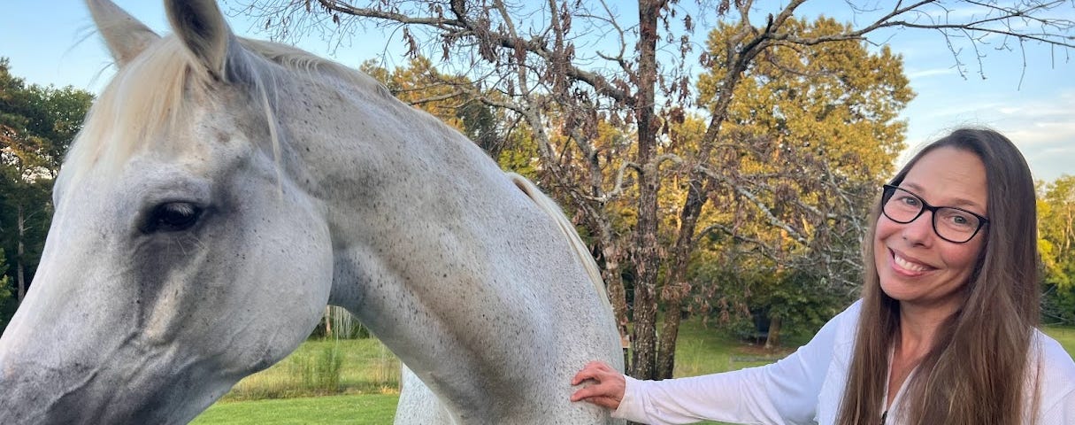 A tall white horse with black spots standing next to a woman with long hair, glasses and a white shirt. The woman’s right hand is on the horse.