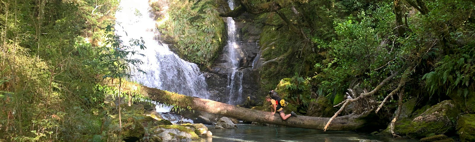Woman wearing a backpack moves while sitting, across a large log over a river with waterfalls in the background. The river is surrounded by large trees.