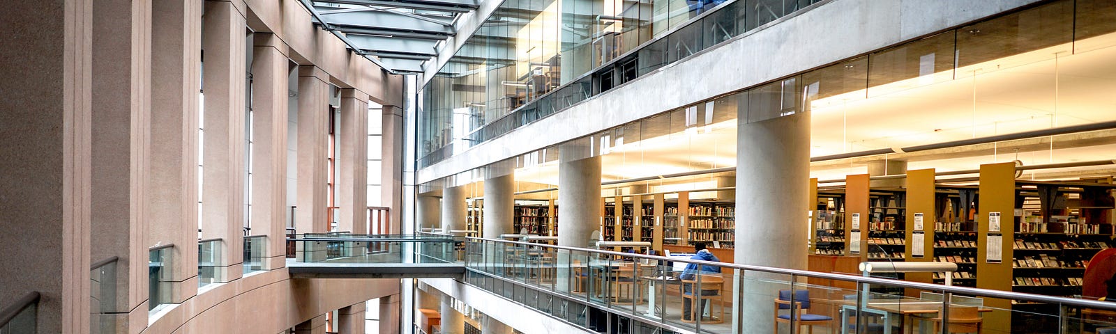 A photo of the Vancouver Library taken from inside. On right side, there are four floors and there are books on the bookshelves and tables and chairs at the end of every floor. On the left side, the wall is curved makes the round shape of the library.