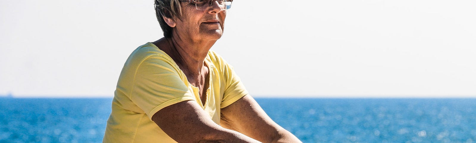 Older woman with glasses sitting on a beach with ocean behind her.