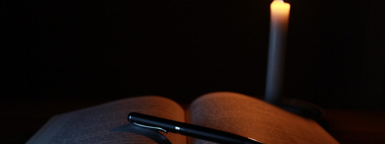 Dark room with one lit candle near an open book with a pen lying on the book diagnally.