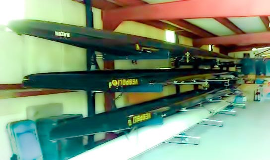 Several Vespoli eight-seater crew shells stored on racks in a boathouse.