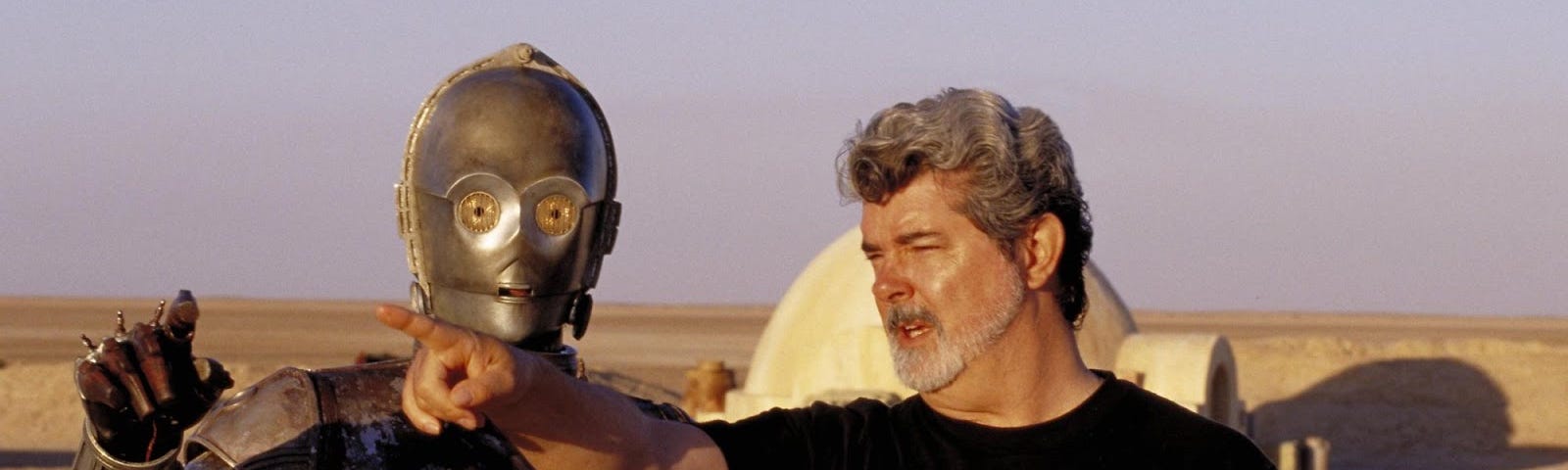 A still from one of the more recent Star Wars movies sets, with the robot C3P0 stood next to George Lucas, who is pointing off screen.