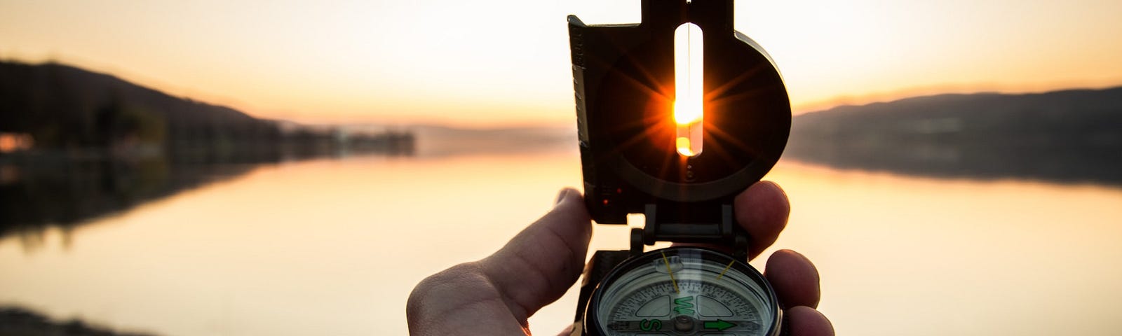 Colour photograph of a person holding a compass up to the horizon overlooking a lake at dawn.