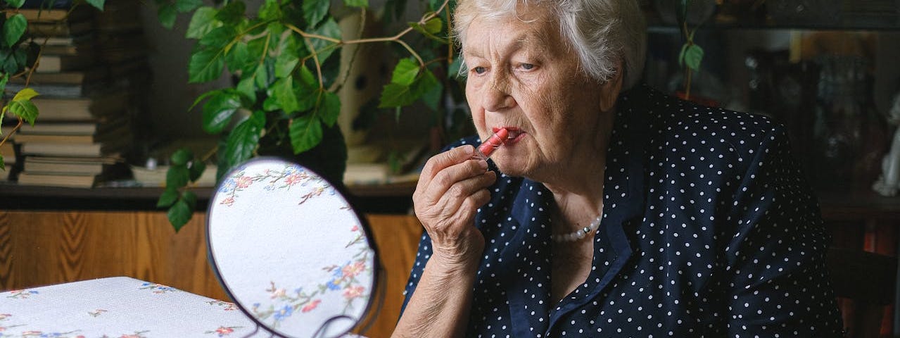 An elderly woman applies lipstick while looking in a small round mirror.