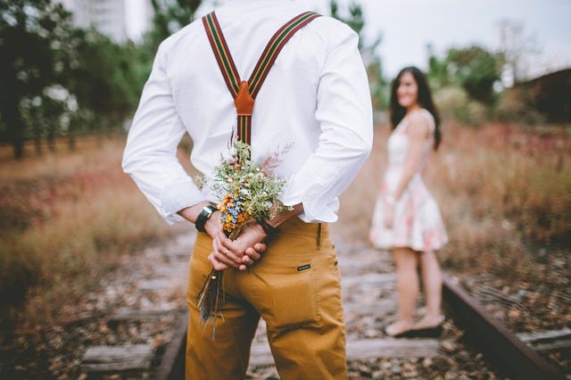 Man with braces, woman in summer dress, man conceals a bunch of flowers behind his back, old railway track, trees