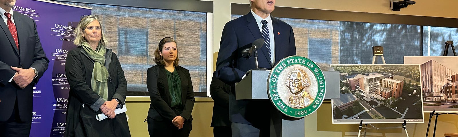 Photo of Gov. Jay Inslee standing at a podium. Behind him are two women and a man as well as some photos showing rendering of what the new hospital will look like when it’s complete.