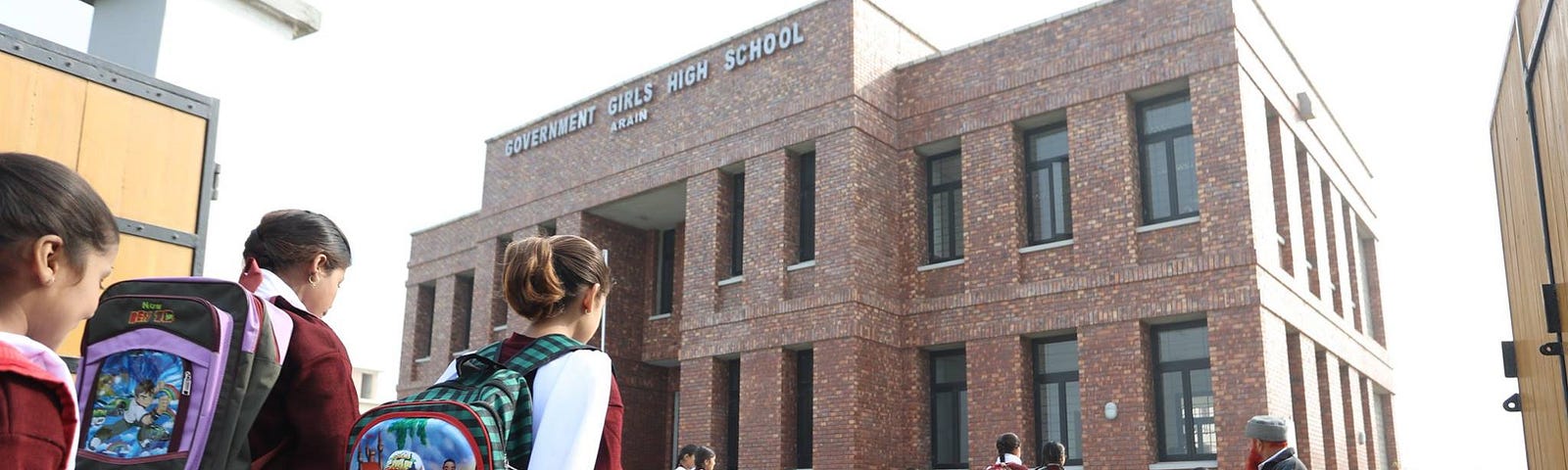 Schoolgirls with backpacks walk towards a newly constructed, two-story brick school. A school official sits outside the school at a desk.