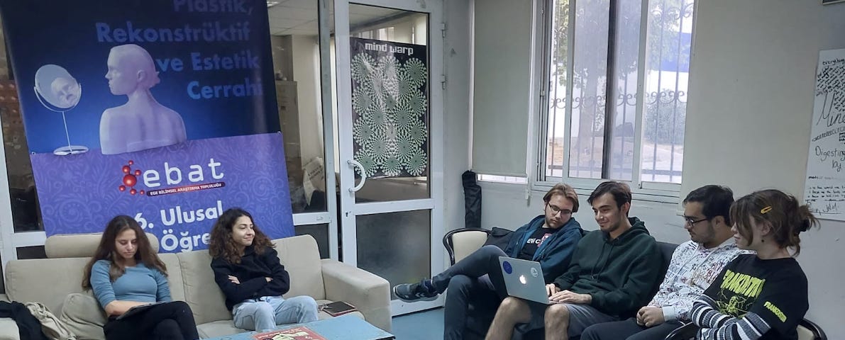 Photo of a medical student lounge with 2 couches and a small table. 2 students sit on the left couch with a poster of the school name behind them. 4 students on the right couch look at a laptop together.