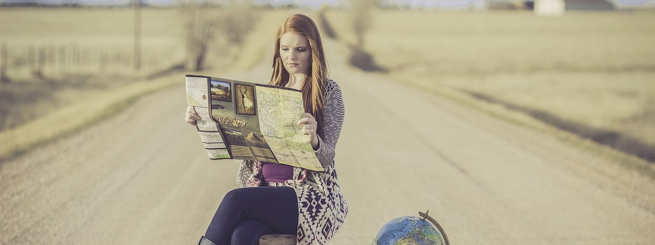 A young woman sitting on a crate in the middle of a country road, open map in hands, a suitcase and globe next to her.