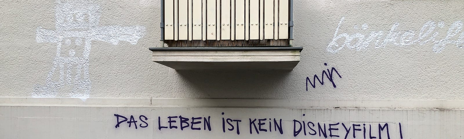 Photo of a beige balcony in Berlin with a beige wall covered in graffiti tags. The one right below the balcony in purple/blue  reads “Das Leben ist Kein Disneyfilm” or Life is not a Disney movie.