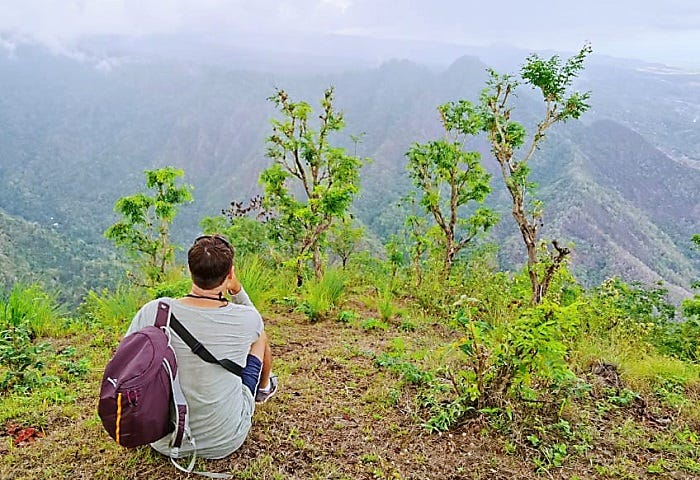 man wearing backpack sitting on hilltop looking at distant valley view