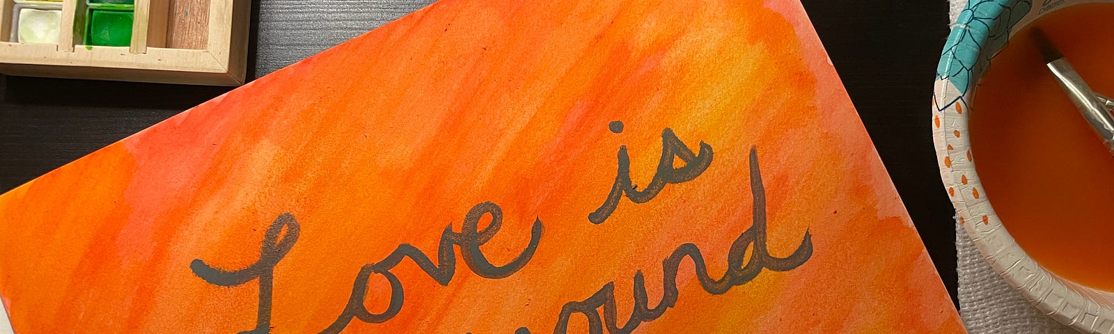 A watercolor painting that says “Love is all around me.” The background colors are red, yellow, and orange. A bowl full of water and a paintbrush are on the right side, atop a paper towel. An open palette of watercolors is to the left.