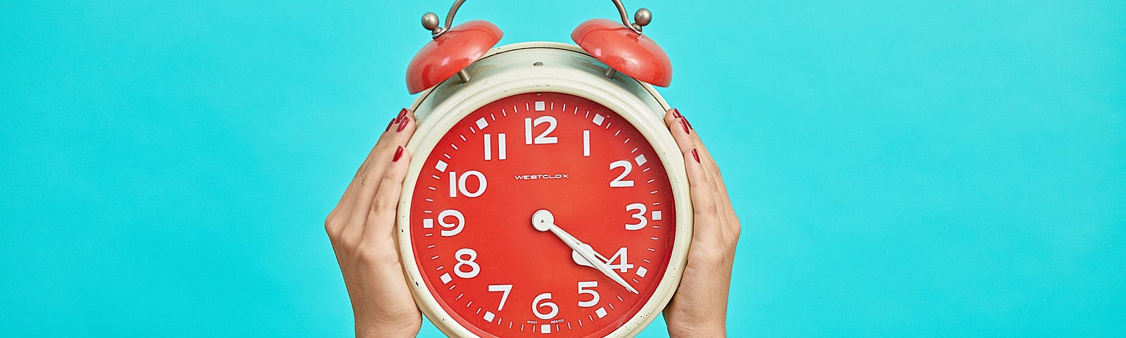 Two hands with red-painted fingernails holding an analog clock with a red face and white numbers and hands