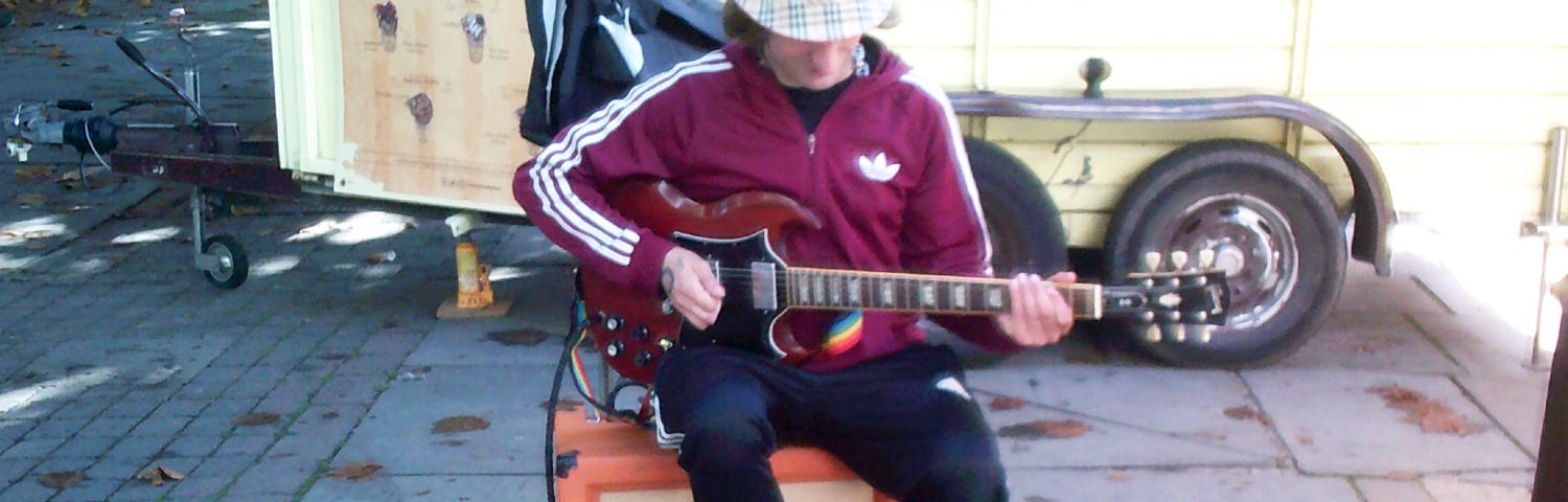 Image of a street busker in a English city market area playing a guitar whilst sat on his amplifier in front of an array of effects pedals