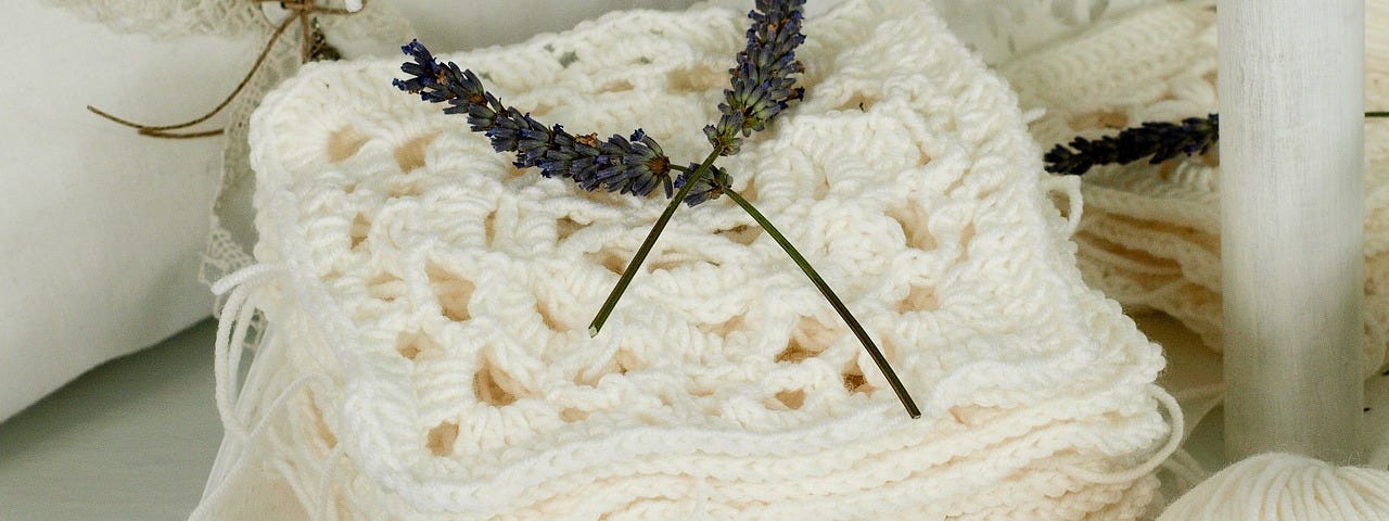 A pile of crocheted granny squares, made from white yarn, with a ball of the same yarn next to the pile. Two sprigs of lavender are crossed on top of the pile.