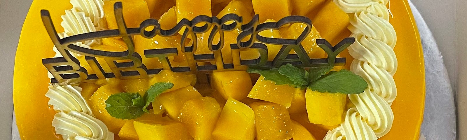 A decorated cake with a mango colored icing and fresh cut mangoes in the middle, surrounded by swirls of vanilla icing. There is a ‘Happy Birthday’ stencil stuck on the cake and the base of the cake says ‘Happy birthday Sameet’