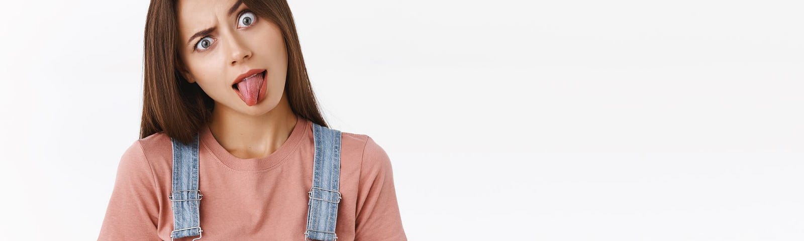 A young woman in denim overalls and a pink tee shirt is sticking out her tongue, and raising one eyebrow as if asking a question.