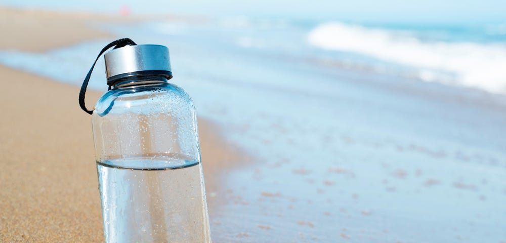 Picture of a reusable bottle with drinking water on the beach, close to the water.