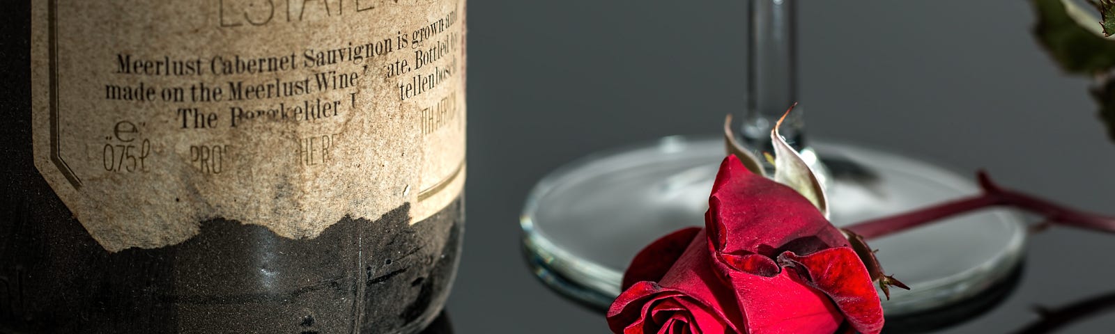 A shiny black surface with a dark red rose laying on it in front of the base of a long stemmed wine glass. The top of the glass is not visible. To the left is the bottom of a wine bottle of cabernet sauvignon, the top of which is also not visible.