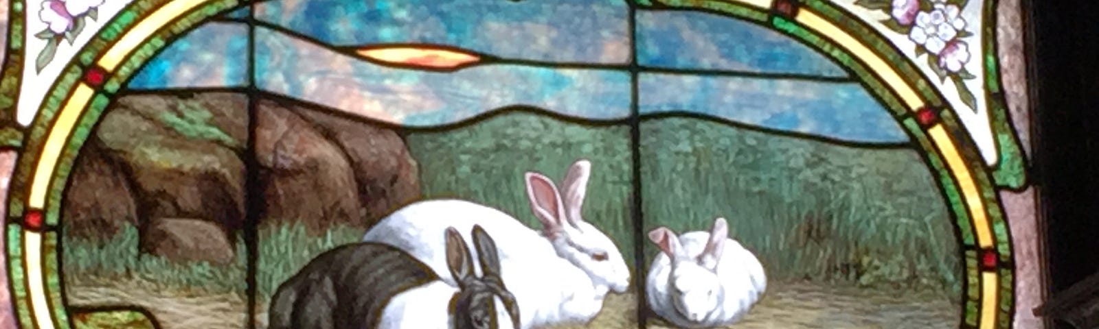 Stained glass window featuring three rabbits — one black and white, and two all-white — with carrots on the ground near them