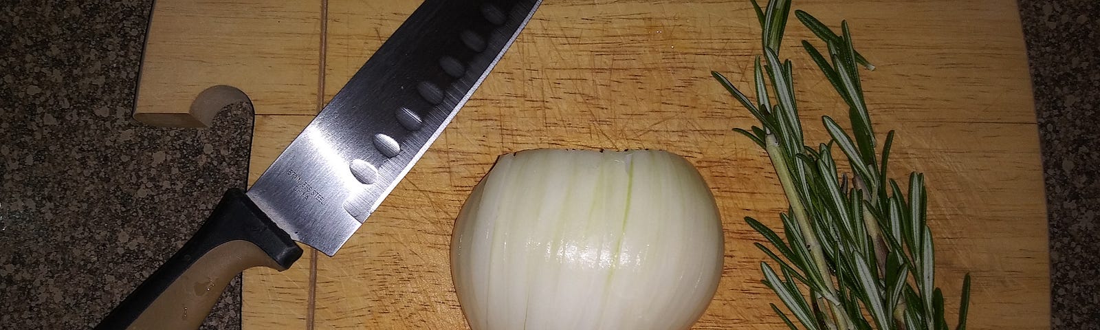 A sharp knife, half of a cut and sliced onion, and two bushy sprigs of fresh rosemary are on a wooden cutting board.