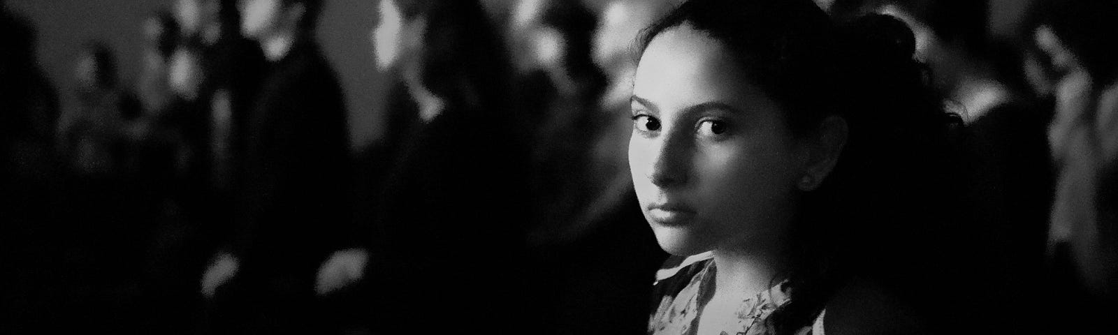 Grayscale picture of crowd with young woman at front, staring into the camera