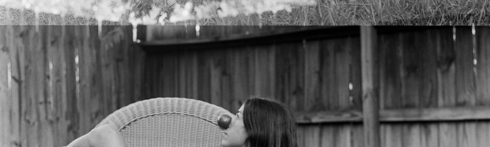 Black and white profile portrait of a woman in a chair wearing a clown nose, outside, legs crossed, fence in the background