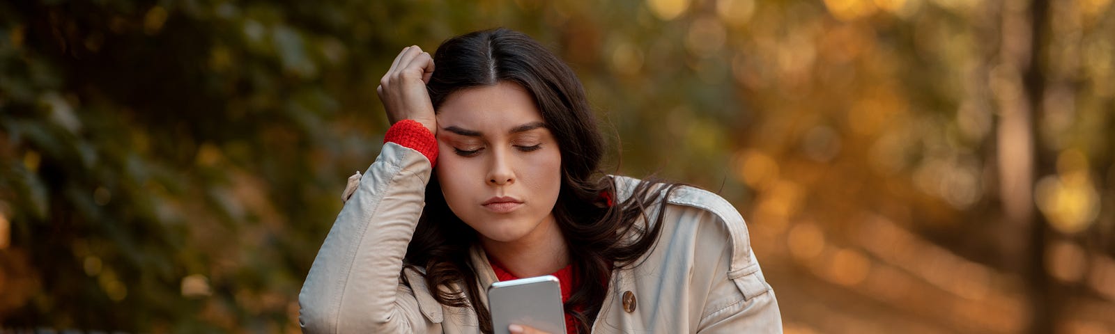 A woman sits on a bench in autumn and grimaces at her phone with a disappointed look.